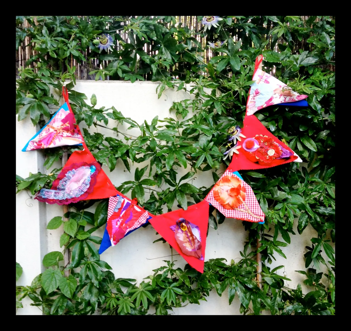 Colorful triangular fabric bunting decorated with patterned patches, hanging on green leafy foliage against a wall